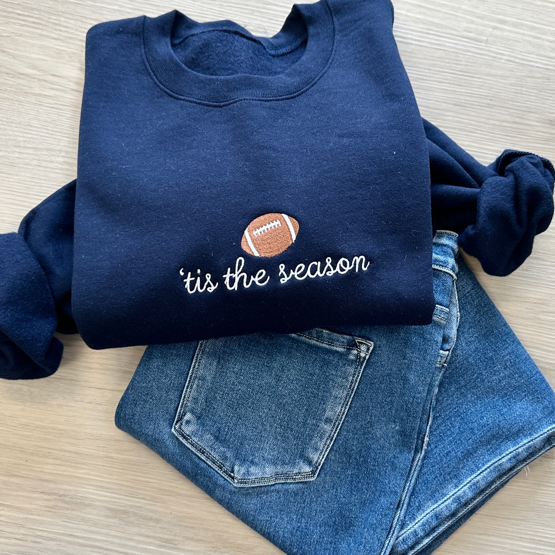 Navy blue sweatshirt with 'tis the season and football icon embroidered on the center chest paired with jeans