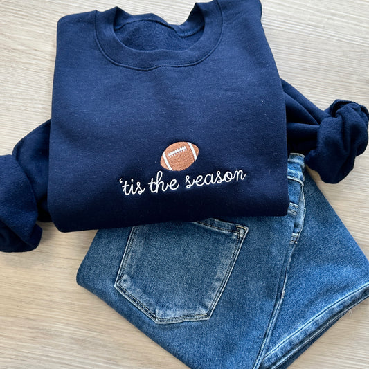 Navy blue sweatshirt with 'tis the season and football icon embroidered on the center chest paired with jeans
