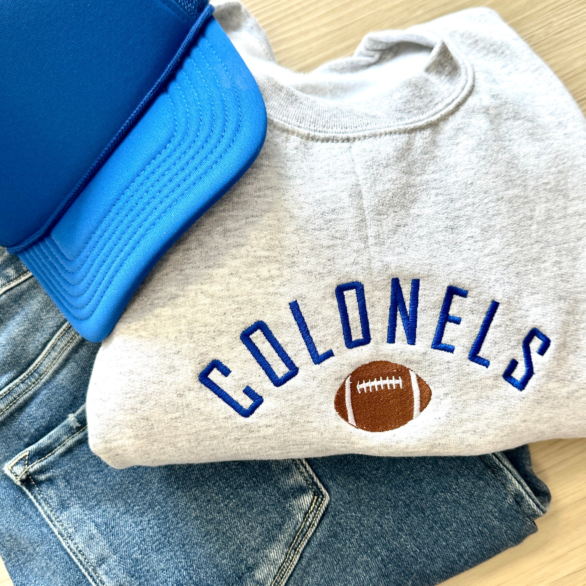 ash sweatshirt with custom embroidery featuring a mascot name in a curved design centered above an embroidered football icon. paired with royal blue foam trucker hat and jeans