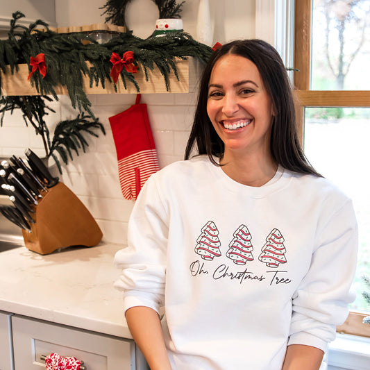 woman sitting in her kitchen wearing a white crewneck sweatshirt with a little debbie christmas tree oh christmas tree printed design