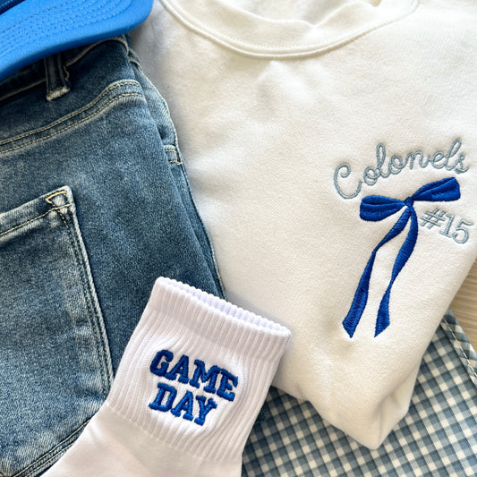 Folded white shirt with 'Colonels #15' and long tail bow embroidery on the left chest, blue cap, and jeans, and game day socks