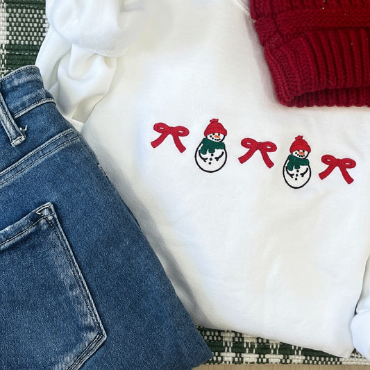 White sweatshirt with red bows and snowmen, blue jeans, and a red beanie on a patterned surface.