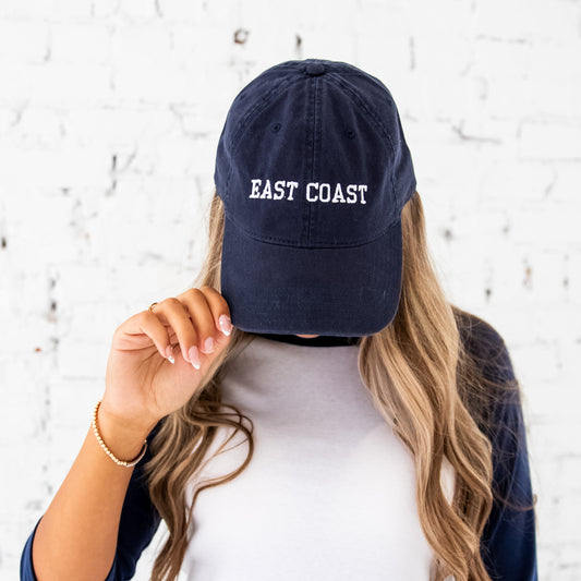 girl with long blonde hair wearing a navy baseball hat with east coast embroidered in an athletic block font and white thread. she's wearing a navy raglan long sleeve top and standing in front of a white brick wall