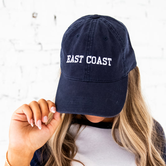 girl with long blonde hair wearing a navy baseball hat with east coast embroidered in an athletic block font and white thread. she's wearing a navy raglan long sleeve top and standing in front of a white brick wall