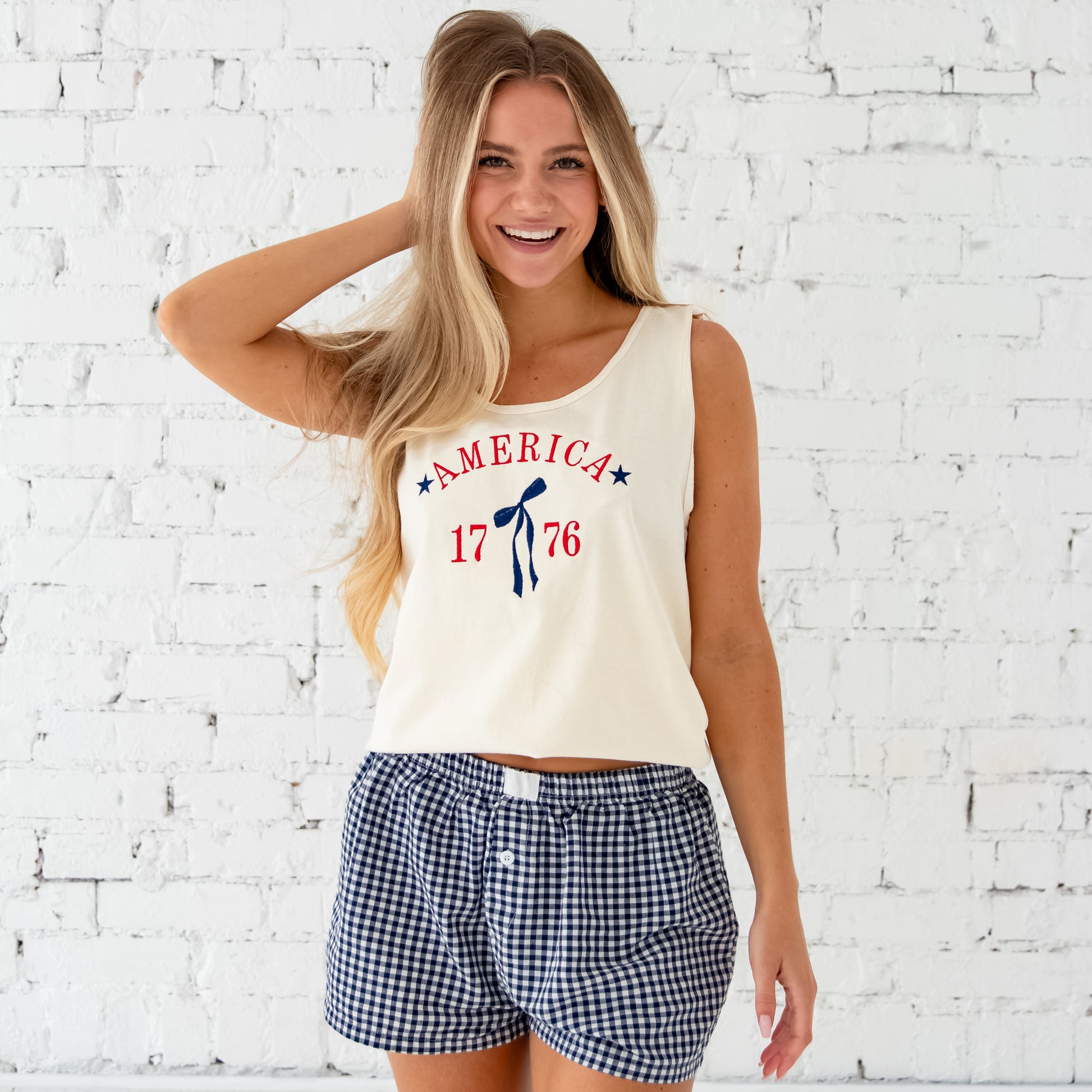 Woman standing in front of a white brick wall wearing an ivory comfort colors tank top and blue and white gingham shorts. Tank top features a custom embroidered design of America arched over a long tail ribbon bow framed by 1776. Embroidery is done in red and navy thread.