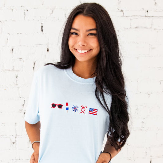 girl standing in front of a white brick wall wearing an oversized comfort colors chambray blue t-shirt with 4th of July inspired embroidered icons: red sunglasses, a red white and blue popsicle, firework, red bow, and an American flag.
