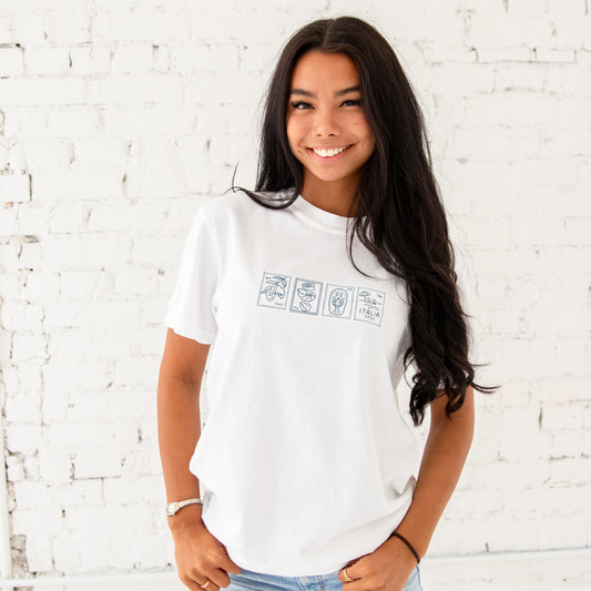 brunette sitting in front of a white brick wall, and she's wearing a white comfort colors t-shirt with a custom Italian stamps outlined embroidered design in blue thread