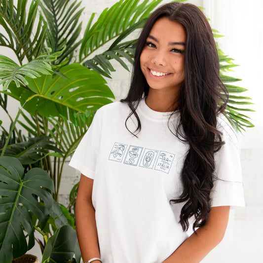 brunette sitting in front of a large palm plant, and she's wearing a white comfort colors t-shirt with a custom Italian stamps outlined embroidered design in blue thread