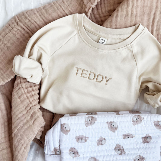 toddler crewneck pullover in natural with a simple, modern uppercase name embroidery in camel thread paired with a quilted teddy bear dopp kit