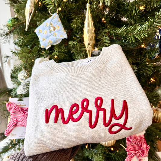 White sweater with 'merry' in red text in front of a Christmas tree.
