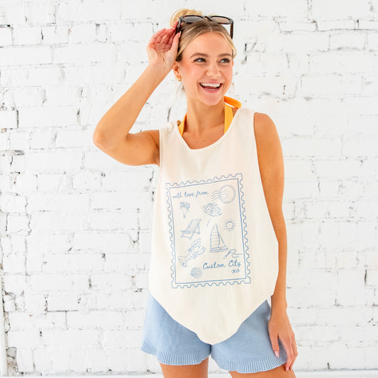 girl standing in front of a white brick wall. Her hair is pulled up into a messy bun with a clip, she has sunglasses resting on top of her head. She's wearing light blue knit shorts and an oversized tank top over an orange bikini. The tank top features a custom printed "With love from" tank top. The design on the tank top has a large stamp outline with fun coastal, beachy icons inside as well as a spot to add a custom city. The tank top is ivory and the print is blue.