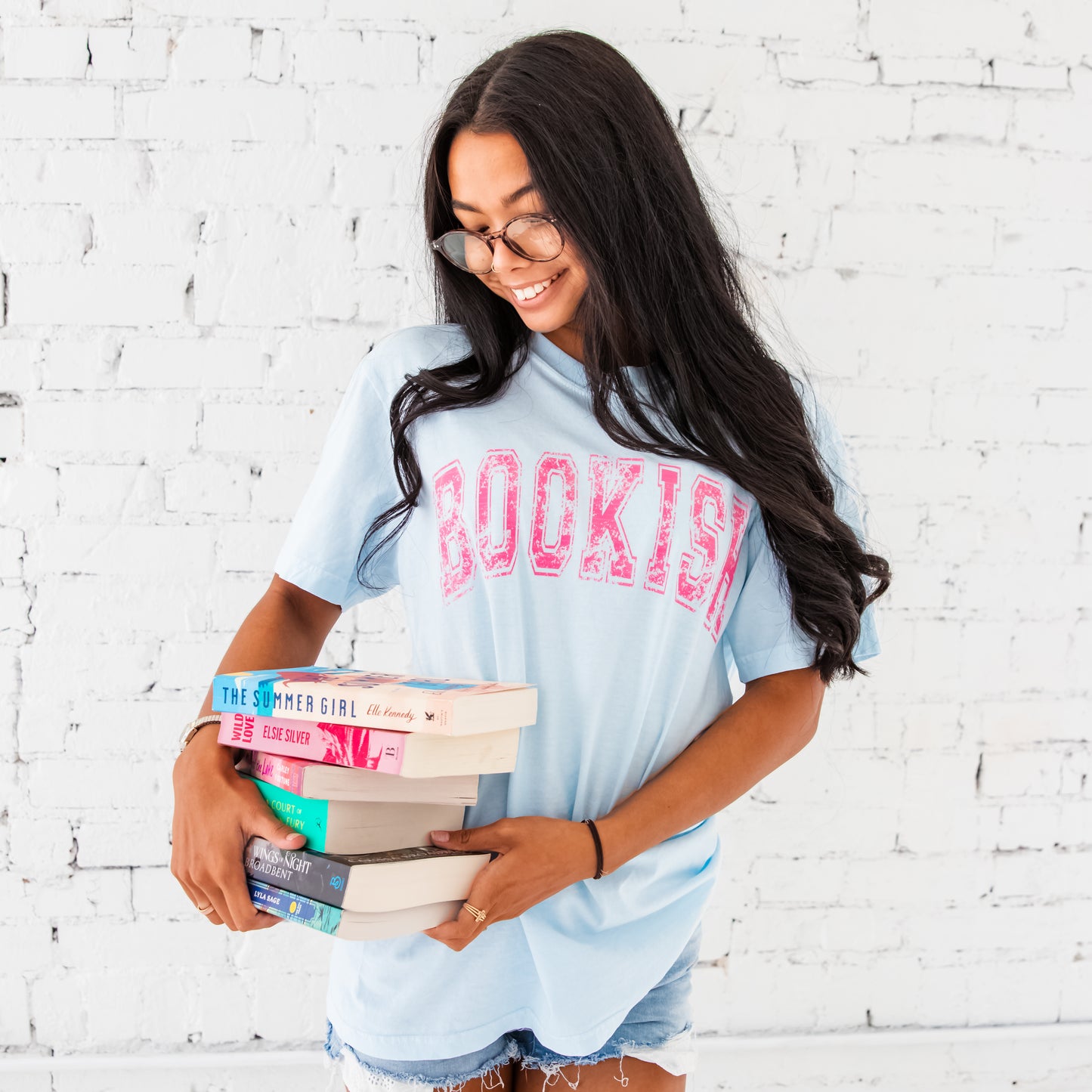 woman wearing chambray shirt with bookish printed across the chest in a distressed vintage athletic font. she is also holding a stack of books