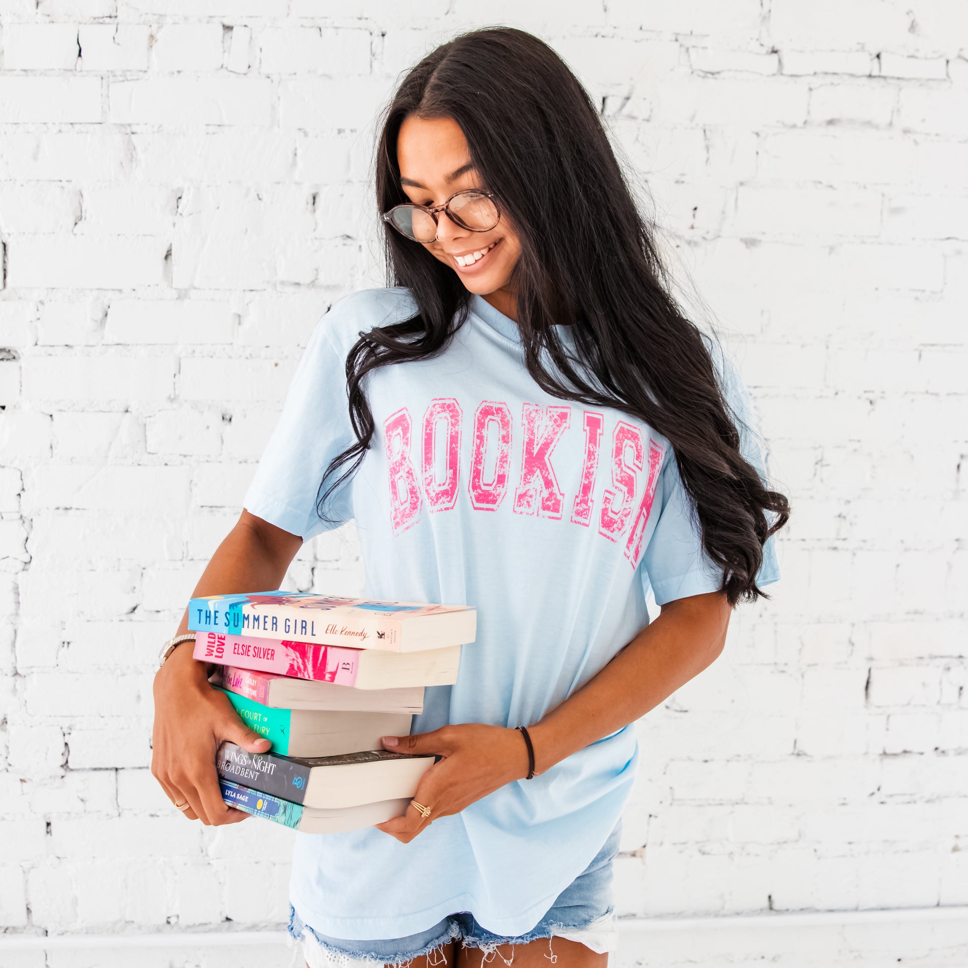 woman wearing chambray shirt with bookish printed across the chest in a distressed vintage athletic font. she is also holding a stack of books