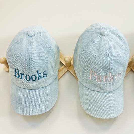 Two light blue baseball caps with embroidered names on a wooden stand.