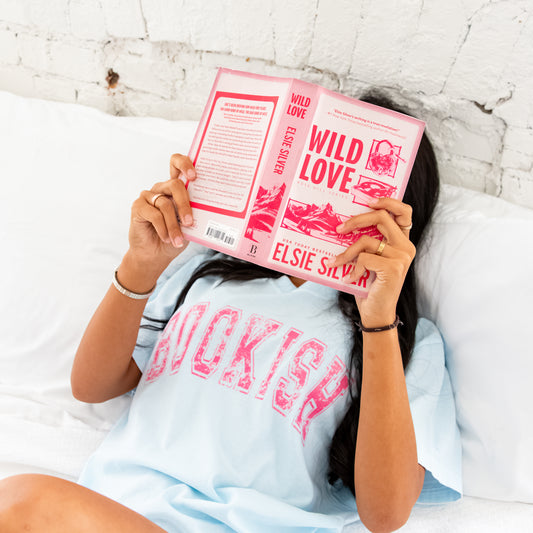 woman laying down, reading a book, wearing chambray comfort colors t-shirt with bookish printed across the chest in a distressed athletic block font and pink ink