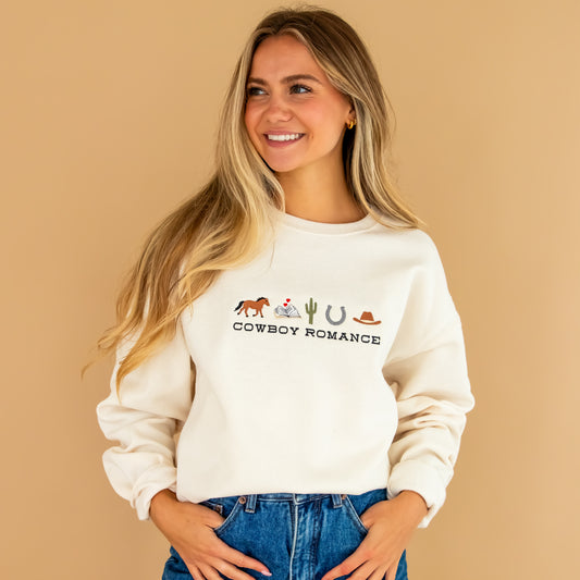 Woman standing wearing sweet cream sweatshirt with brown horse, book, cactus, horse shoe, cowboy hat, and cowboy romance embroidered across the chest, partnered with denim shorts.