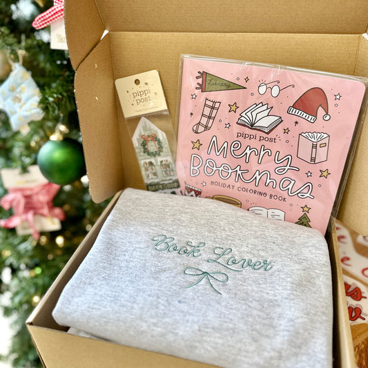 Box with 'Book Lover' sweatshirt and 'Merry Bookmas' coloring book and bookmark in front of a Christmas tree.