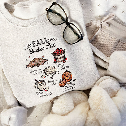 heather oat crewneck sweatshirt with a cute stitch fall bucket list inspired design featuring a pile of leaves, basket of apples, a baked apple pie, up of cider, and carved pumpkins