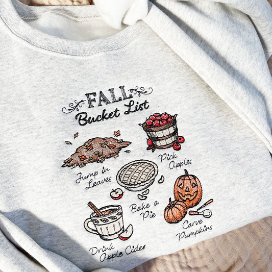 heather oat crewneck sweatshirt with a cute stitch fall bucket list inspired design featuring a pile of leaves, basket of apples, a baked apple pie, up of cider, and carved pumpkins