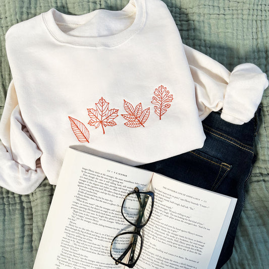 styled flat lay of a sweet cream crewneck sweatshirt with embroidered outlines stitched fall leaves in a pumpkin spice thread color with jeans, a book, and glasses