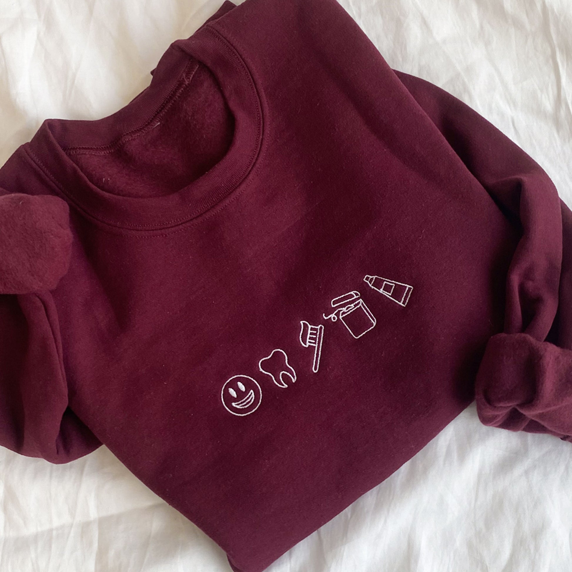 maroon crewneck sweatshirt with 5 dental icons embroidered across the chest in white thread. the icons include in order from right to left a simlie face, outline tooth, toothbrush, floss, and tooth paste tube.