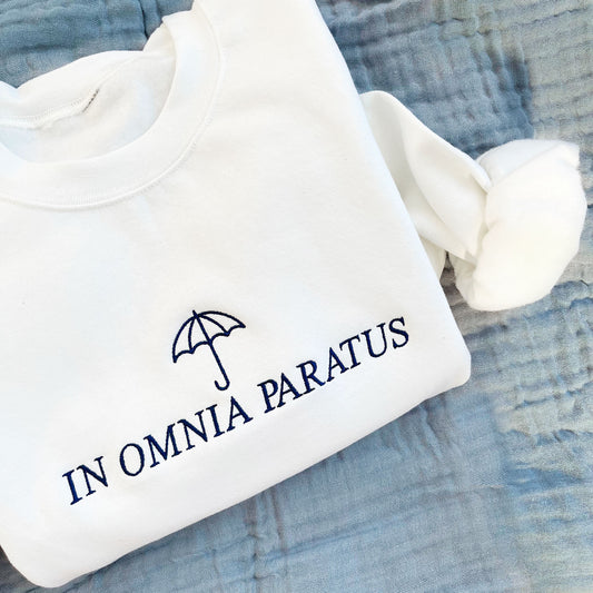 flat lay of a white crewneck sweatshirt with embroidered umbrella and "IN OMNIA PARATUS" gilmore girls saying in navy thread across the chest.