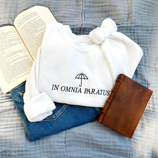 flat lay of a white crewneck sweatshirt with embroidered umbrella and "IN OMNIA PARATUS" gilmore girls saying in navy thread across the chest.