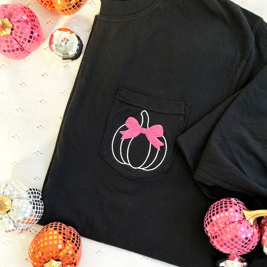 styled fall flat lay of a long sleeve black comfort colors pocket tee with embroidered white pumpkin and pink bow design on the pocket