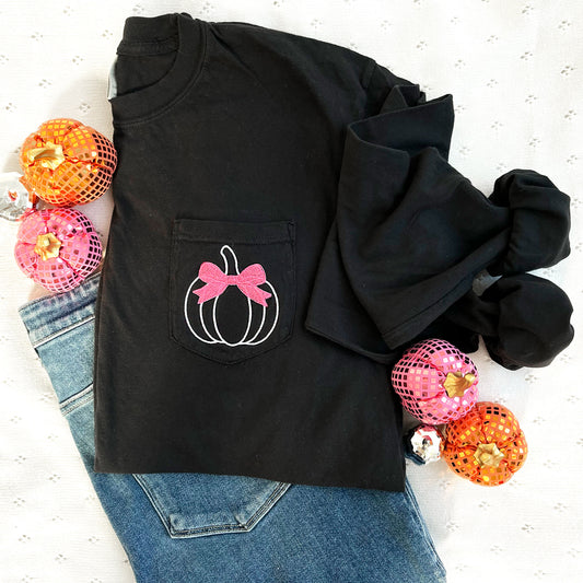 styled fall flat lay of a long sleeve black comfort colors pocket tee with embroidered white pumpkin and pink bow design on the pocket