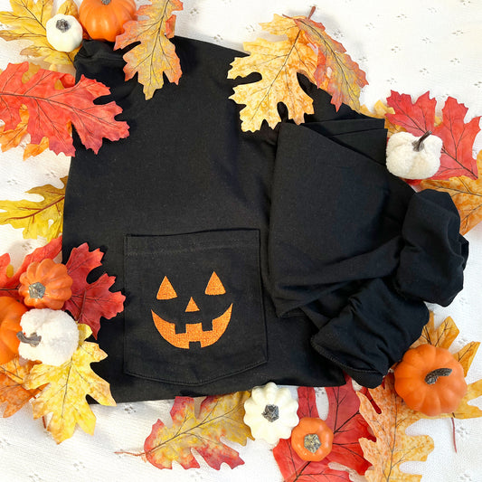 styled fall flat lay of a long sleeve black comfort colors pocket tee with embroidered jack-o-lantern face on the pocket in pumpkin spice thread.