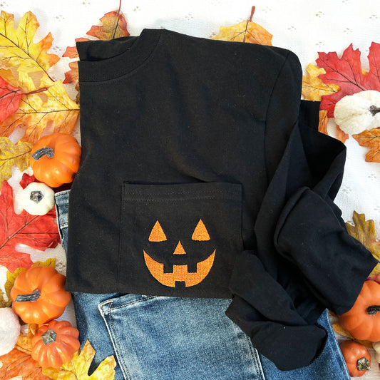 styled fall flat lay of a long sleeve black comfort colors pocket tee with embroidered jack-o-lantern face on the pocket in pumpkin spice thread.