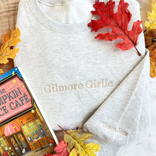 styled fall flat lay of  of an ash crewneck sweatshirt with embroidered  Gilmore Girlie across the chest and Team Jess embroidered on the sleeve both in camel thread.