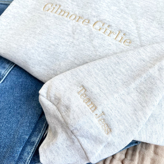 close up of an ash crewneck sweatshirt with Team Jess embroidered on the sleeve in camel thread.