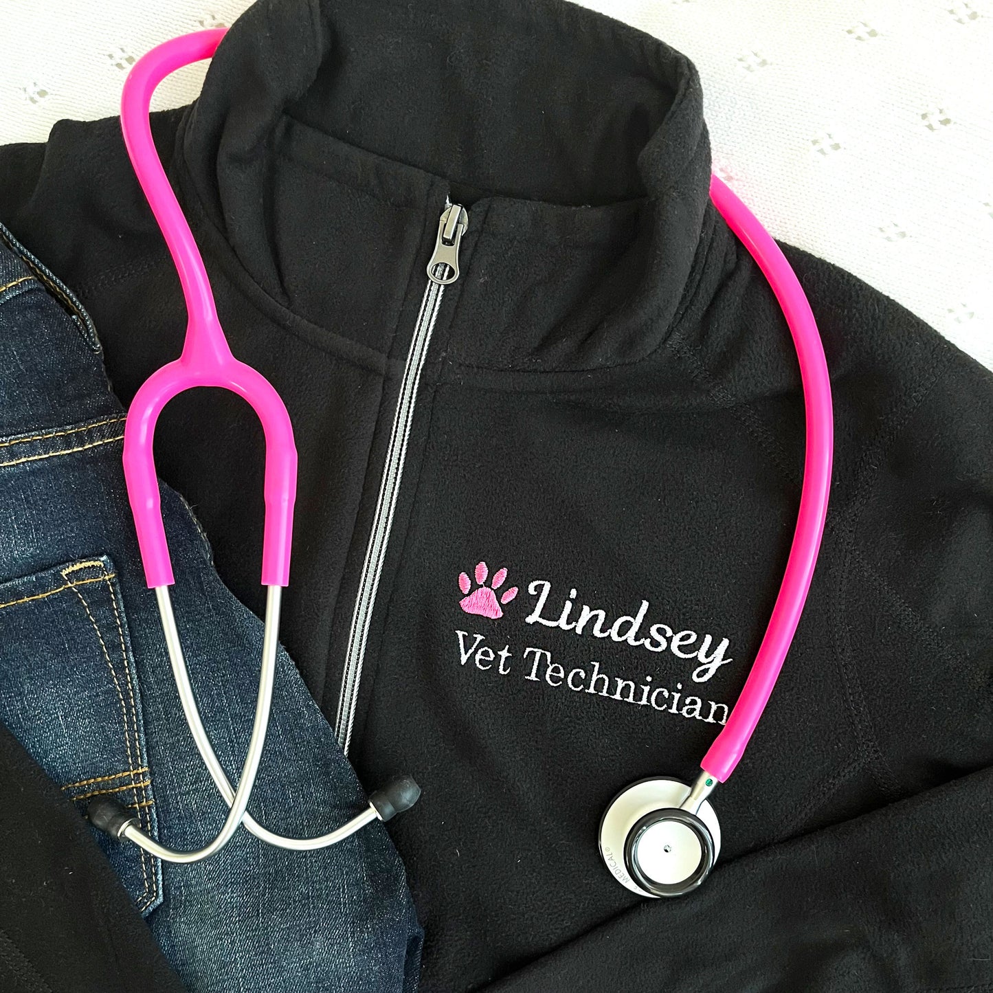 flat lay of a black full zip fleece jacket with embroidered pink paw print and name Lindsey with Vet Technician embroidered on the left chest in white thread
