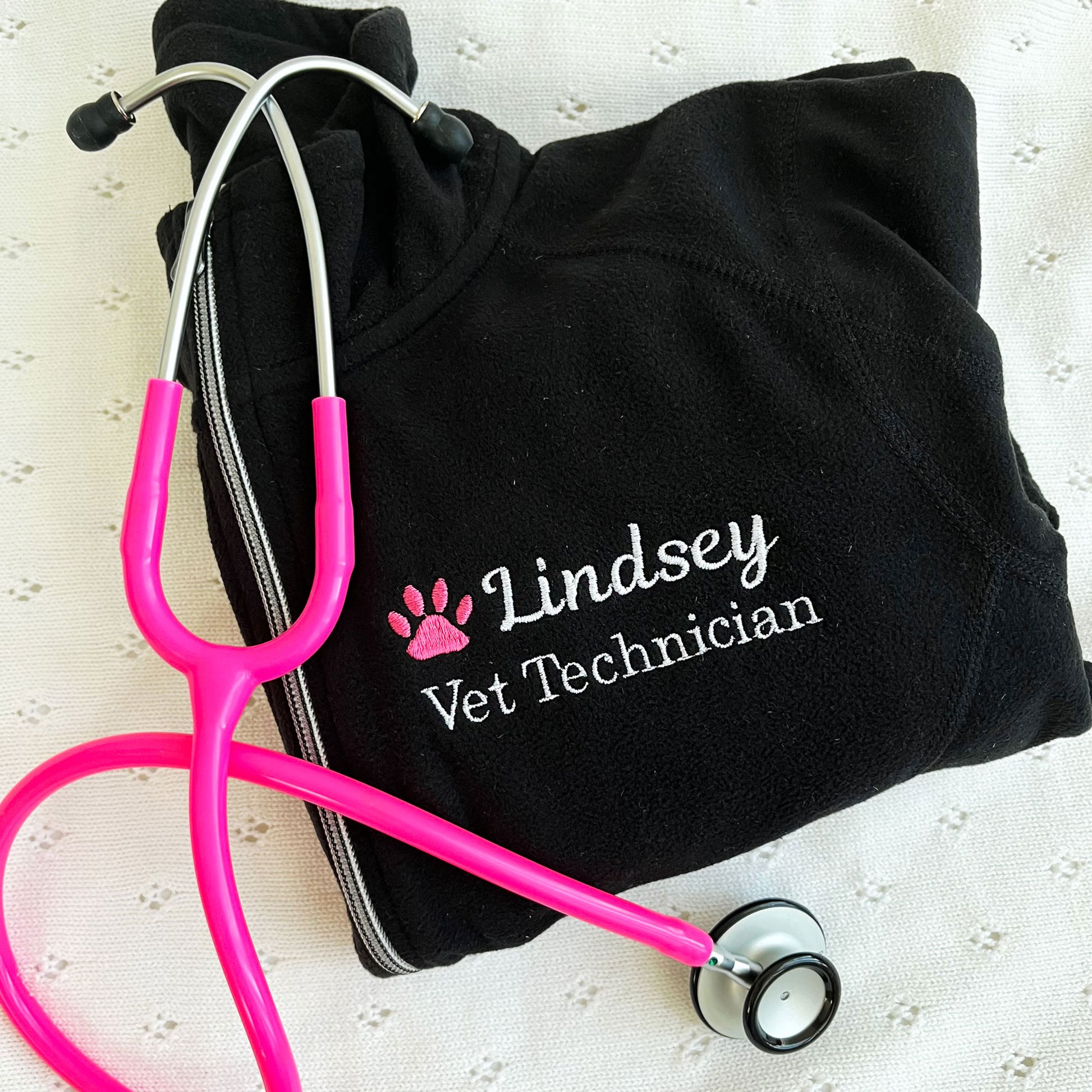 flat lay of a black full zip fleece jacket with embroidered pink paw print and name Lindsey with Vet Technician embroidered on the left chest in white thread
