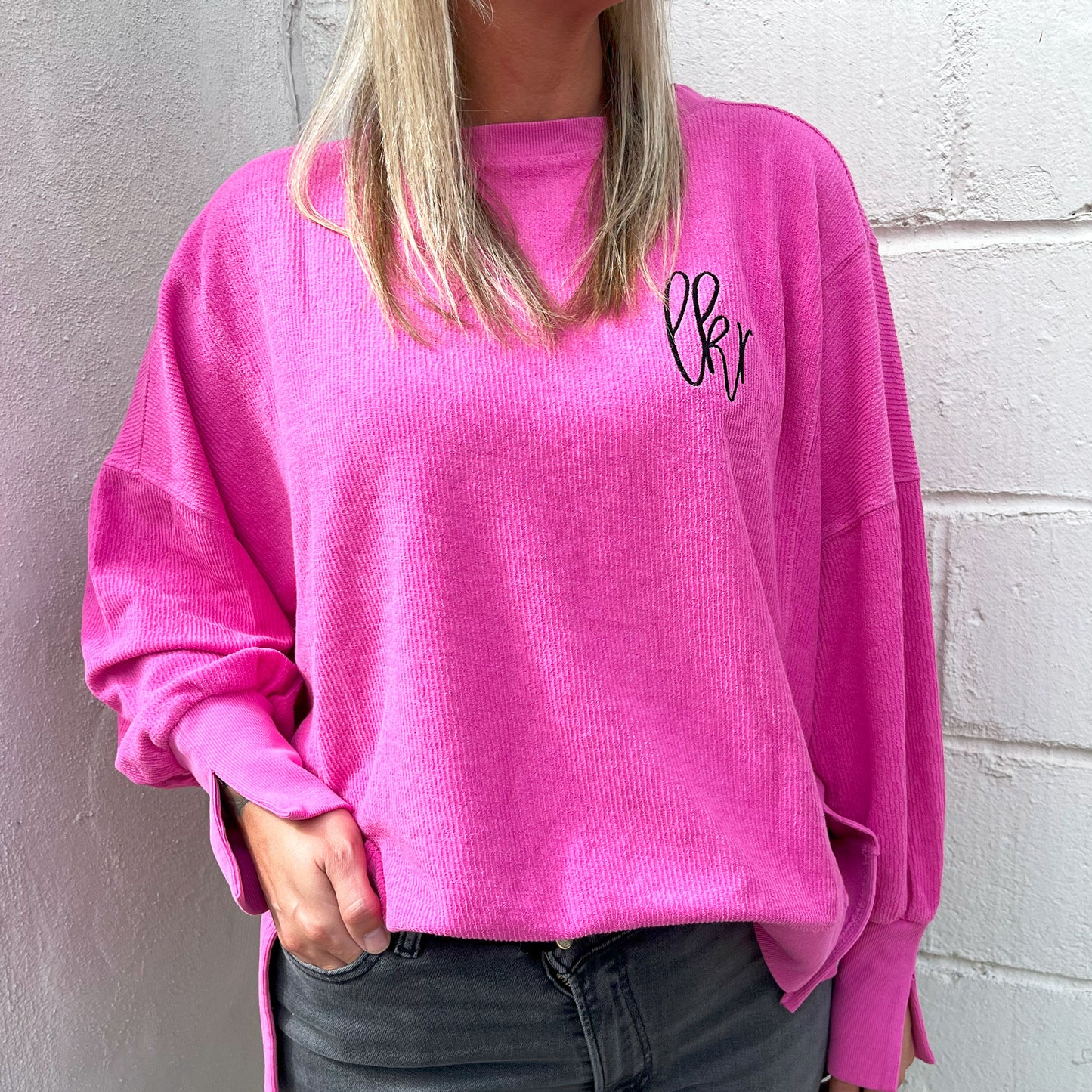 Woman wearing an oversized boxy cropped ribbed crewneck sweatshirt with split hem and cuff details in the pinkberry color. on the left chest is an embroidered monogram in black thread.