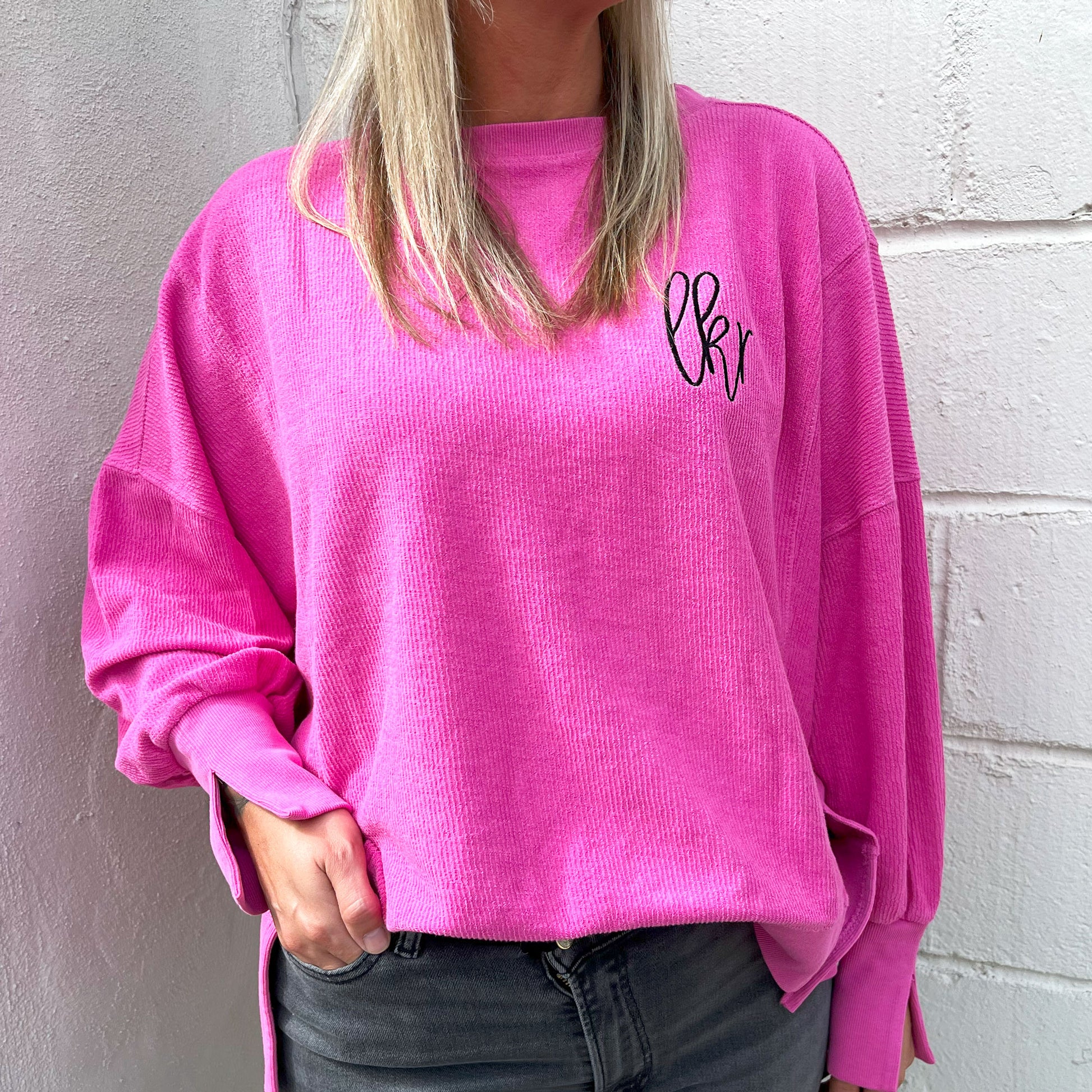 Woman wearing an oversized boxy cropped ribbed crewneck sweatshirt with split hem and cuff details in the pinkberry color. on the left chest is an embroidered monogram in black thread.