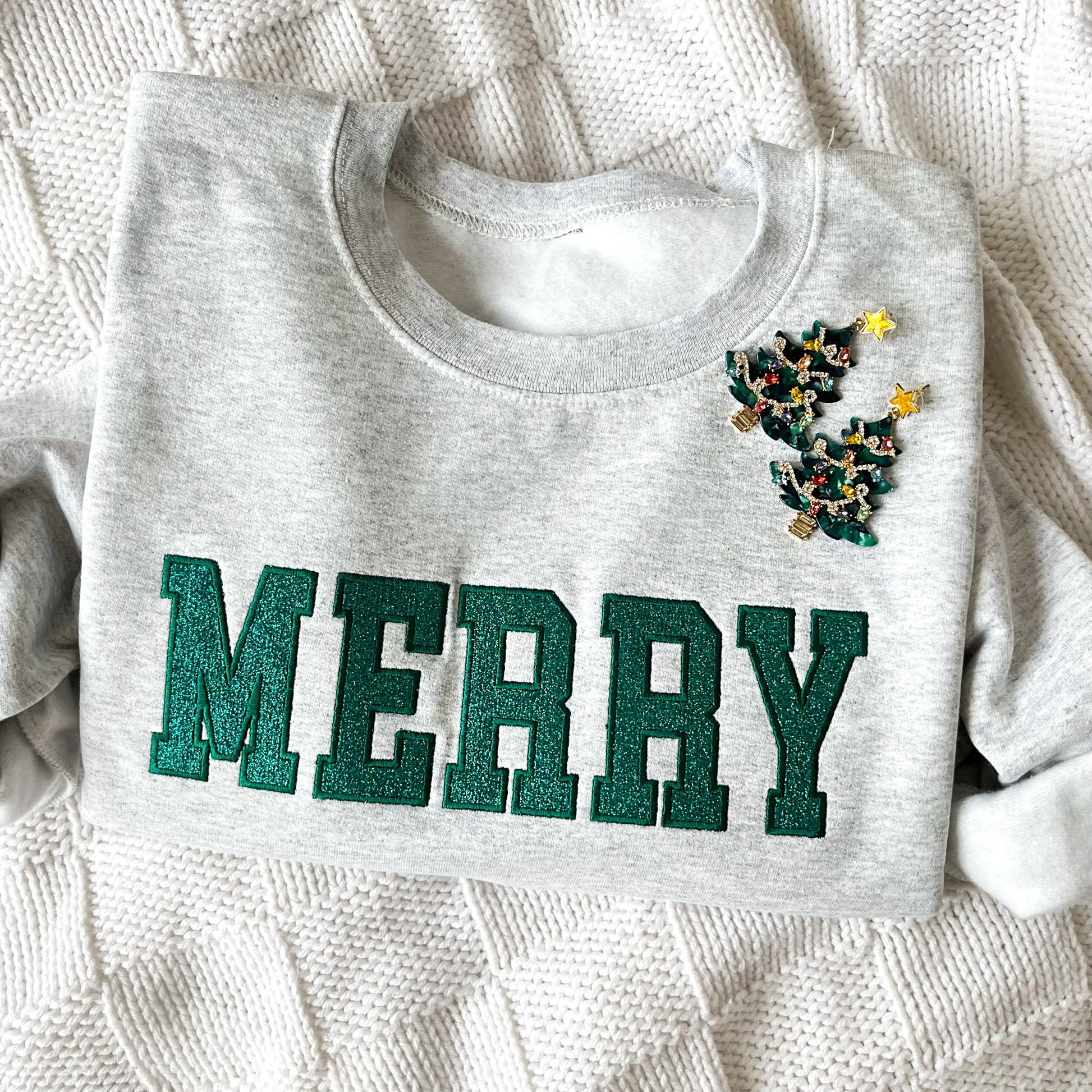 close up of a flat lay of an ash crewneck sweatshirt with embroidered outline merry in all caps and green thread. In the outline embroidered text is a glittery green fill. The sweatshirt is styled with Christmas tree earrings