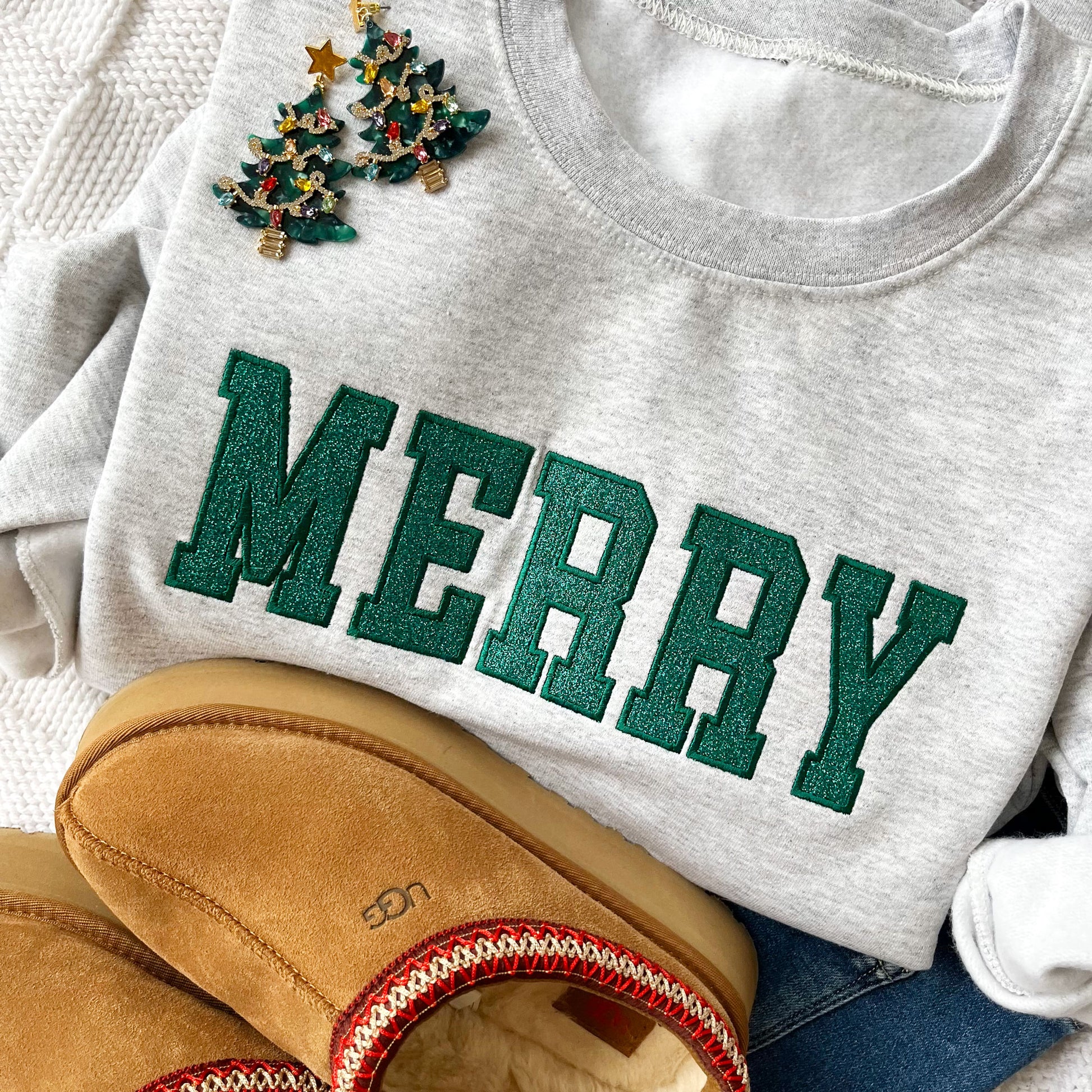 close up of a flat lay of an ash crewneck sweatshirt with embroidered outline merry in all caps and green thread. In the outline embroidered text is a glittery green fill. The sweatshirt is styled with Christmas tree earrings and ugg slippers