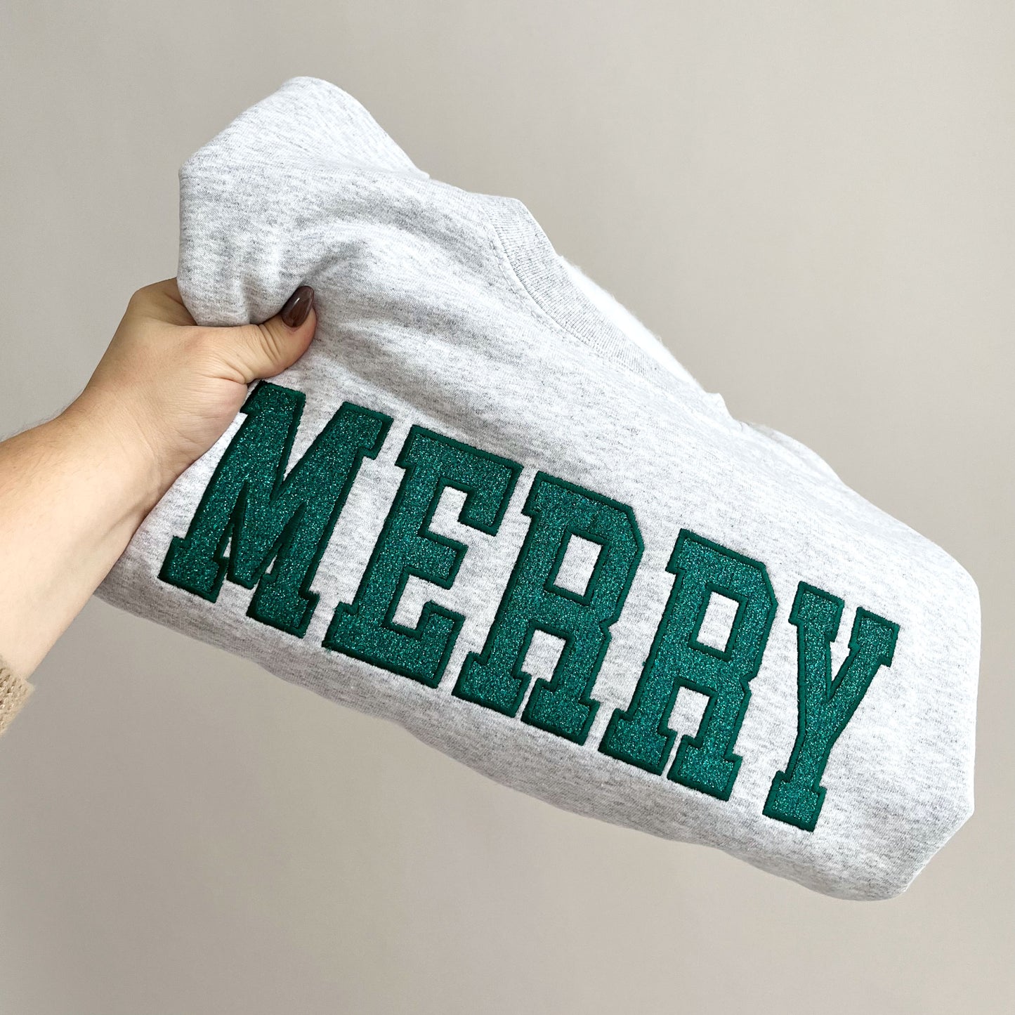 an ash crewneck sweatshirt with embroidered outline merry in all caps and green thread. In the outline embroidered text is a glittery green fill.