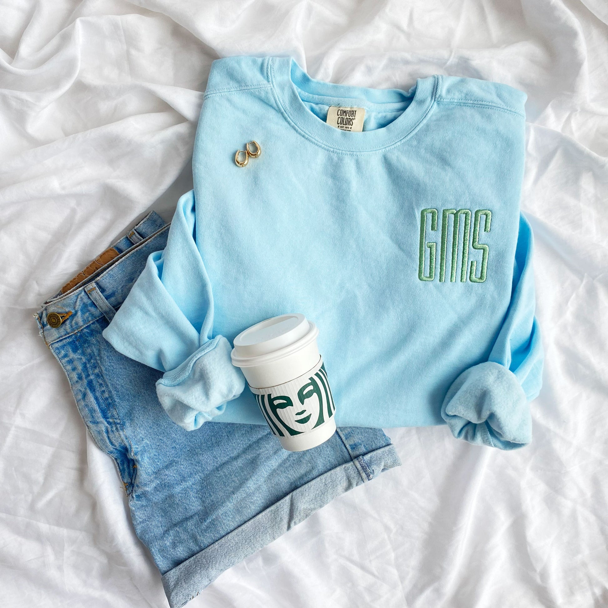Chambray comfort colors crewneck sweatshirt with GMS embroidered monogram in silver sage thread styled with jean shorts, jewelry, and a Starbucks cup
