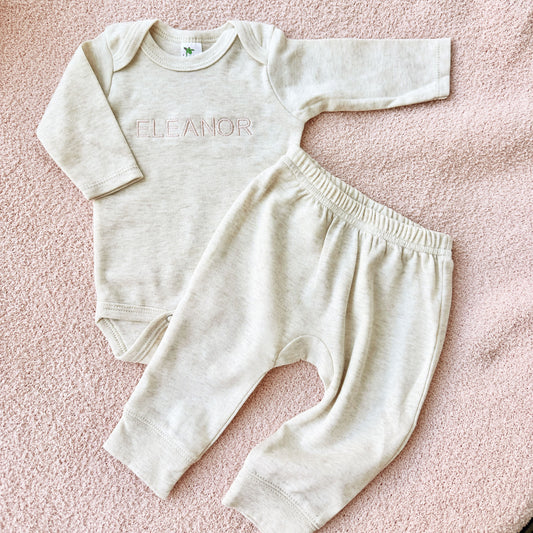 heather oatmeal infant long sleeve bodysuit with custom name embroidered in powder pink thread in Arial uppercase font across the center chest paired with heather oatmeal joggers