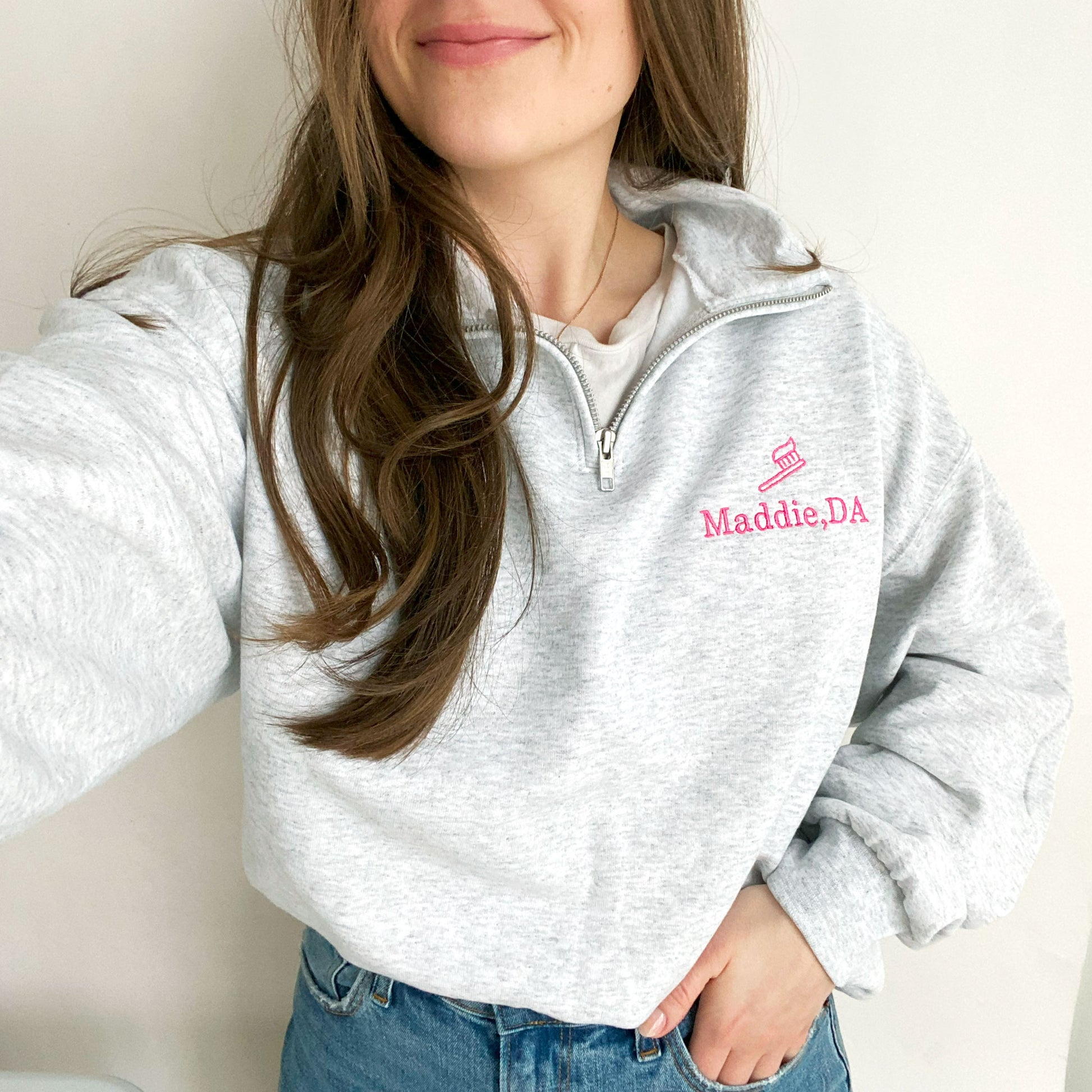 young woman wearing an ash quarterzip sweatshirt with pink embroidered tooth brush and name Maddie, DA on the left chest