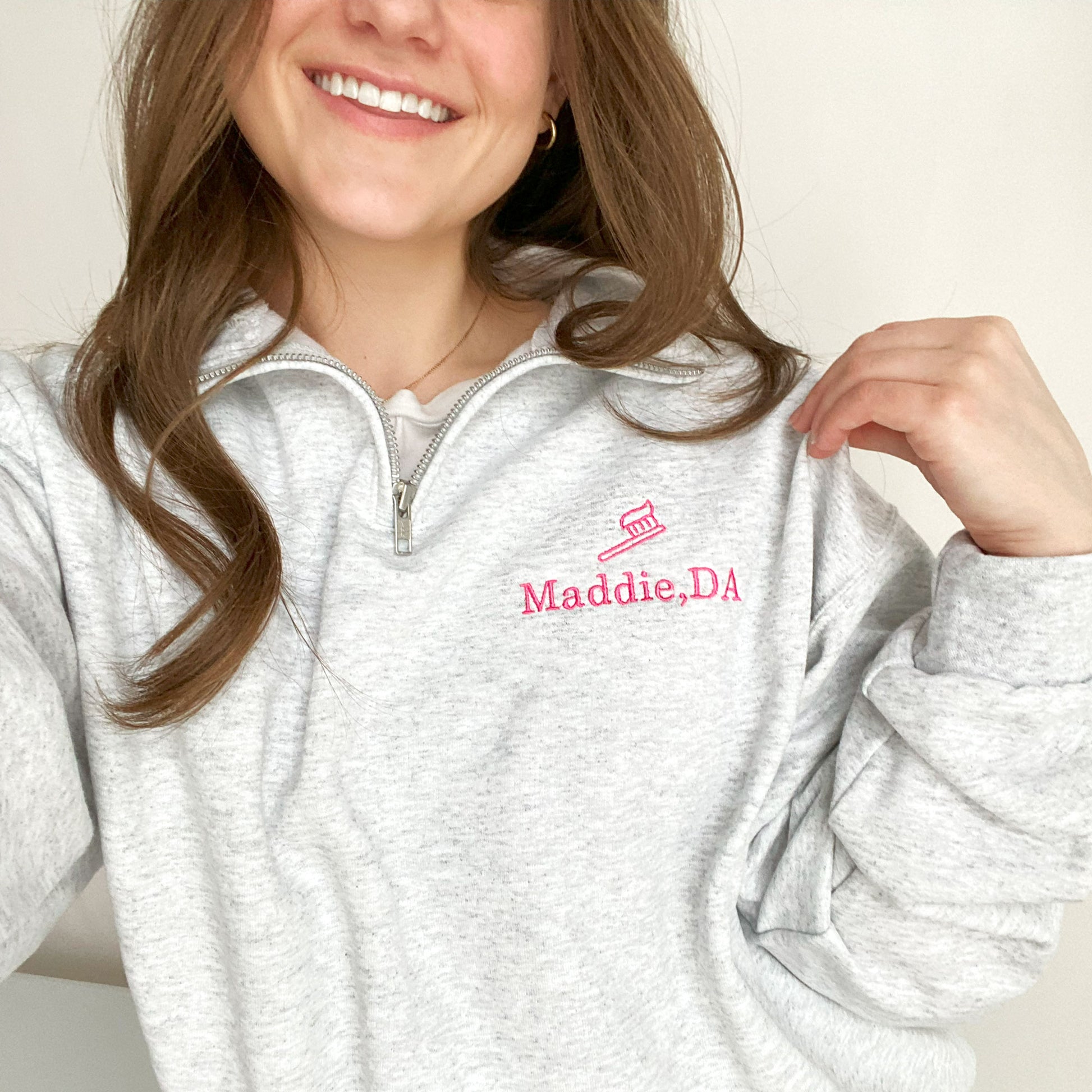 young woman wearing an ash quarterzip sweatshirt with pink embroidered tooth brush and name Maddie, DA on the left chest