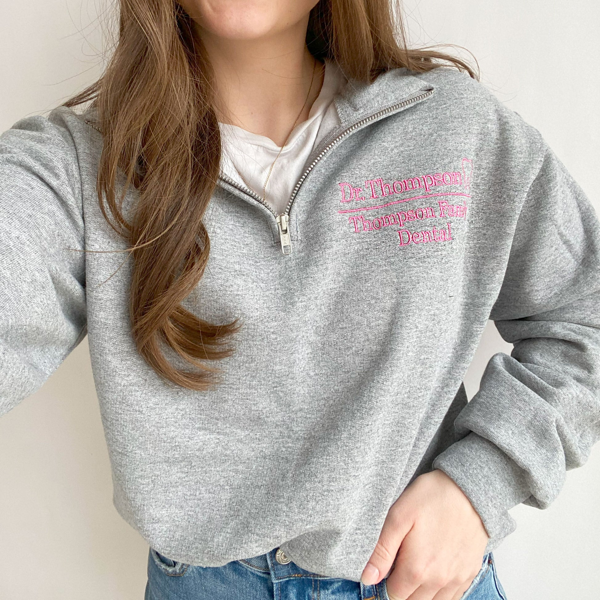 young woman wearing an oxford quarter zip sweatshirt. On the left chest in embroidered Dr. Thompson Thompson Family Dental and mini outline tooth in pink thread