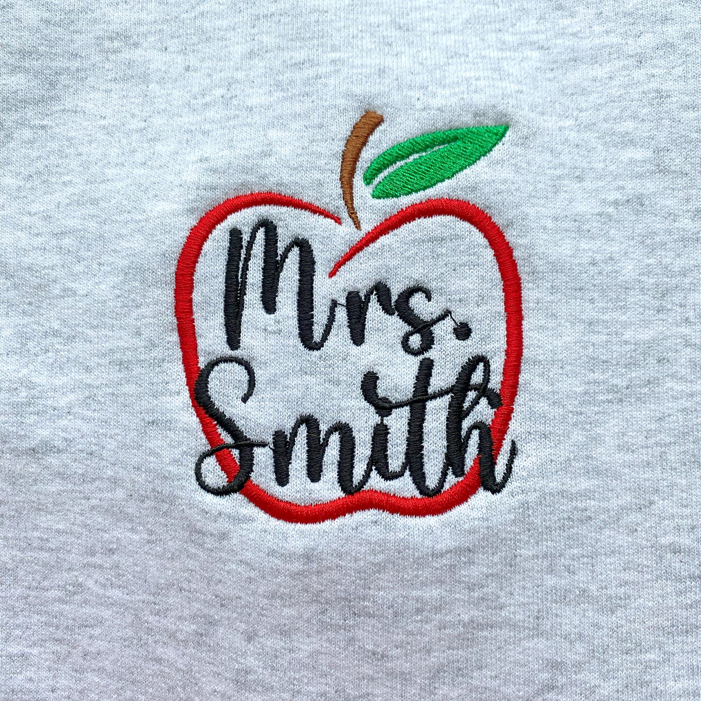 close up of an ash crewneck sweatshirt showcasing the apple outline embroidered design and name mrs smith inside the apple in black thread
