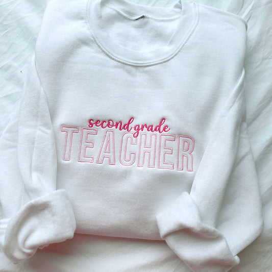 flat lay of a white crewneck sweatshirt with custom grade level and teacher embroidered design in pink and baby pink threads