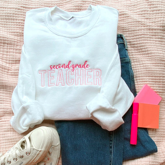flat lay of a white crewneck sweatshirt with custom grade level and teacher embroidered design in pink and baby pink threads