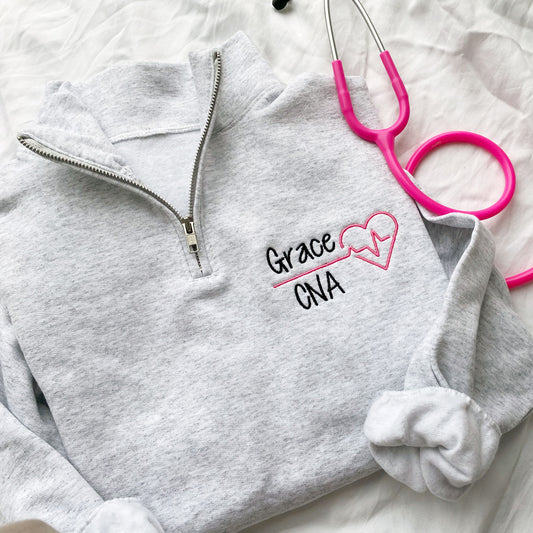 up close flat lay of an Ash quarterzip sweatshirt with custom nurse heart and heartbeat design in pink and name in credentials in black thread