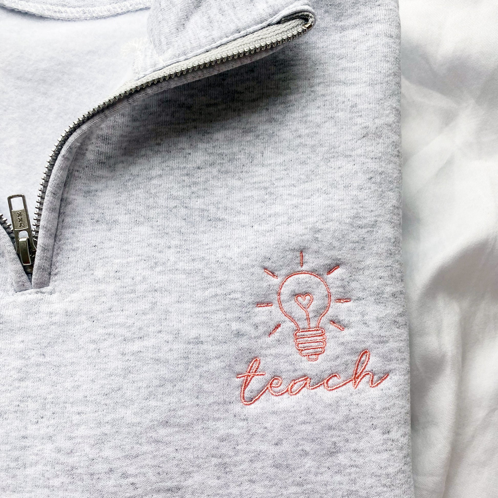 close up of an embroidered design of a lightbulb with 'teach' underneath on a light grey quarter zip sweatshirt